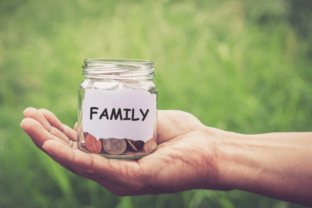 A hand holds a glass jar filled with coins, labelled "FAMILY" on a piece of paper, symbolising family savings. The green, blurry background suggests an outdoor setting.