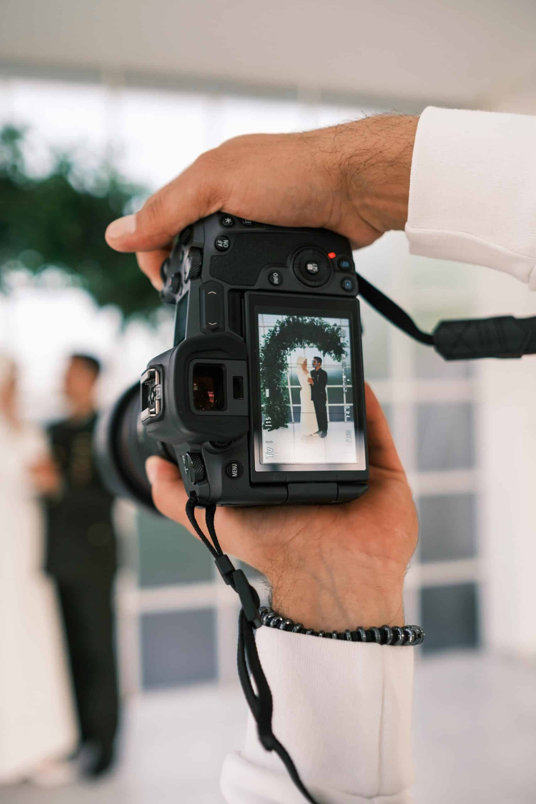 A close-up of a camera held by a person, showing its display screen with a bride and groom standing under an arch, blurred in the background—capturing a moment cherished by couples and wedding suppliers alike.