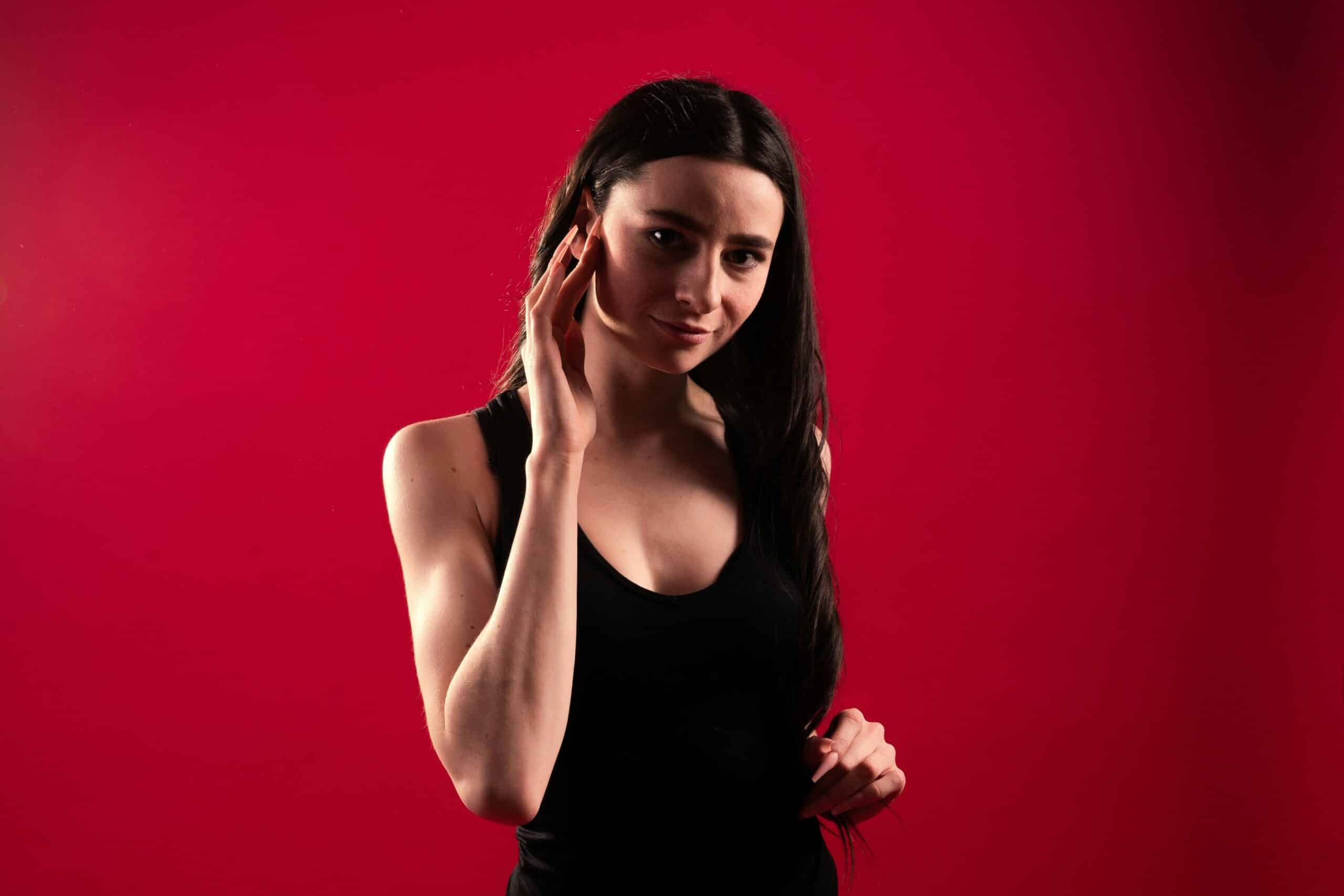 A woman with a striking appearance, long dark hair, and a black sleeveless top stands against a bright red background, touching her face with one hand and looking at the camera with a slight smile.
