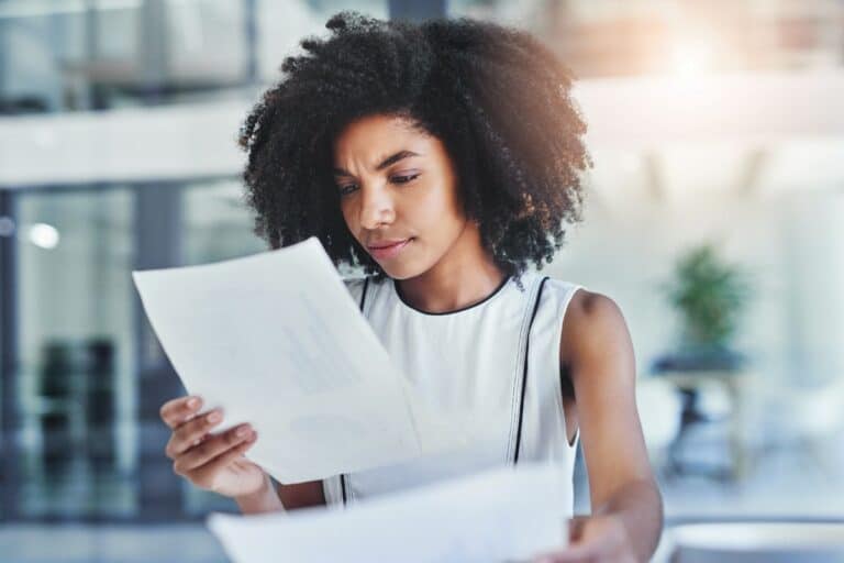 A woman with curly hair and a sleeveless top stands indoors, appearing like a private investigator as she closely examines two sheets of paper. She looks focused and thoughtful, with blurred modern office elements in the background.