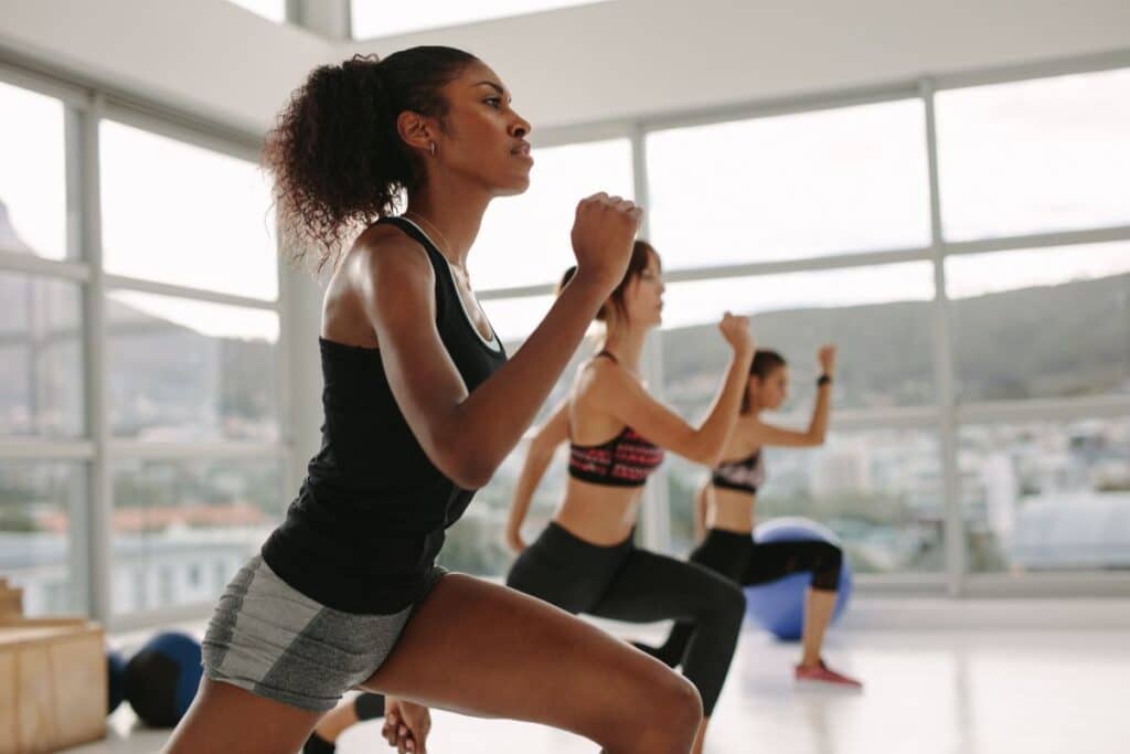 Three women exercise in a bright fitness studio, performing lunges in sportswear. Large windows reveal a cityscape as natural light fills the room, creating an inspiring fitness atmosphere.