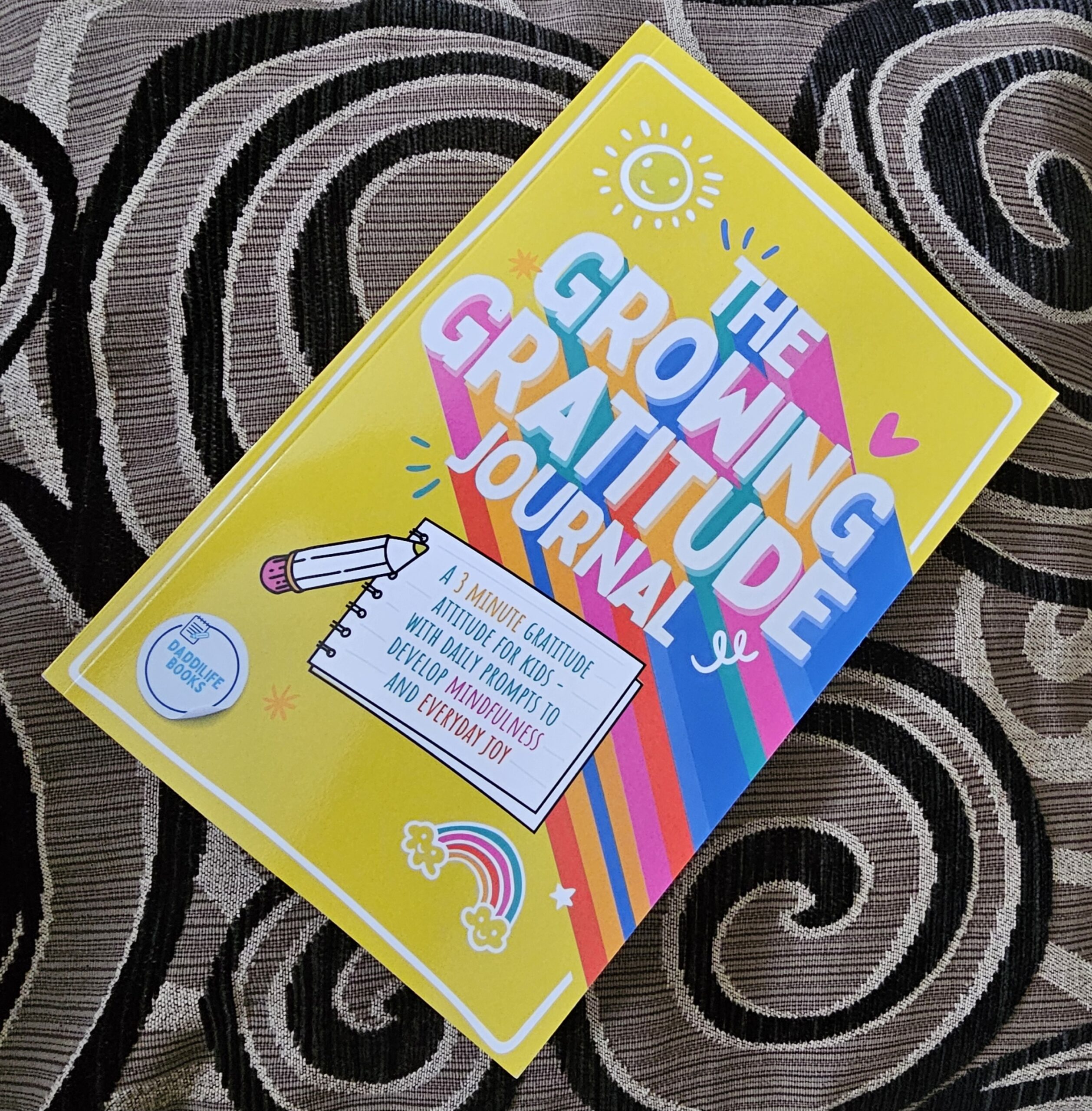 A colourful gratitude journal titled "The Growing Gratitude Journal," decorated with rainbows, stars, and playful doodles, rests on a patterned fabric background.
