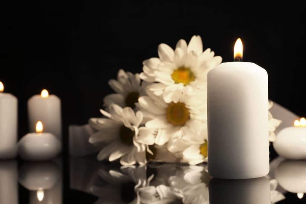 A cluster of white daisies is arranged behind several lit white candles on a reflective surface, with a black background creating a calm and serene atmosphere—perfect for displaying treasured Ashes Jewellery.