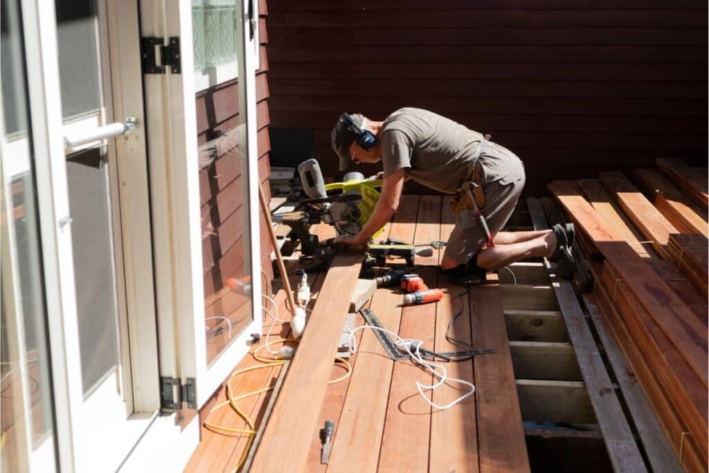 A person wearing headphones and work gear kneels on a sunlit wooden decking, using power tools to install planks for a home project. Various tools and cables are scattered around the busy work area.