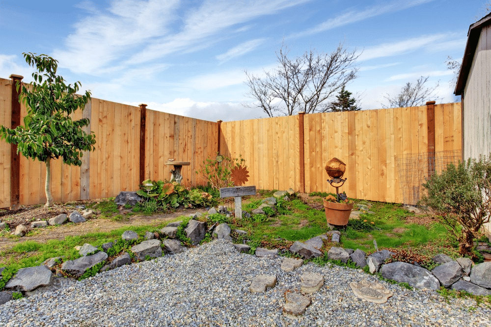 A small back garden with gravel paths, large rocks, and various plants is enclosed by tall wooden fencing. A potted sculpture, birdbath, and decorative metal sunflower are visible under a partly cloudy sky.