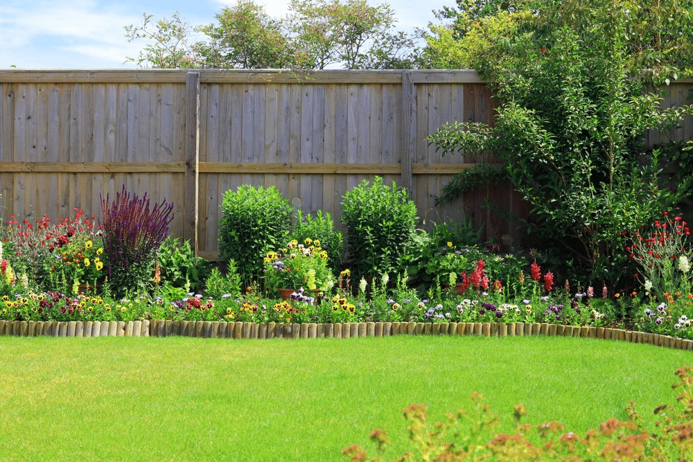A neatly maintained back garden with wooden fencing, lush green grass, and a colourful border of blooming flowers and bushes along the fence. Trees and shrubs provide additional greenery in the background.