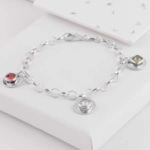 A silver chain Ashes Jewellery bracelet with three gemstone charms—one red, one yellow, and one clear—displayed on a white surface with a gift box in the background.