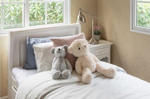 A neatly made, sleep-friendly bedroom features white and pastel bed linen, two teddy bears side by side on top, and a bedside table with a lamp and small plant beside a window showcasing lush green foliage outside.