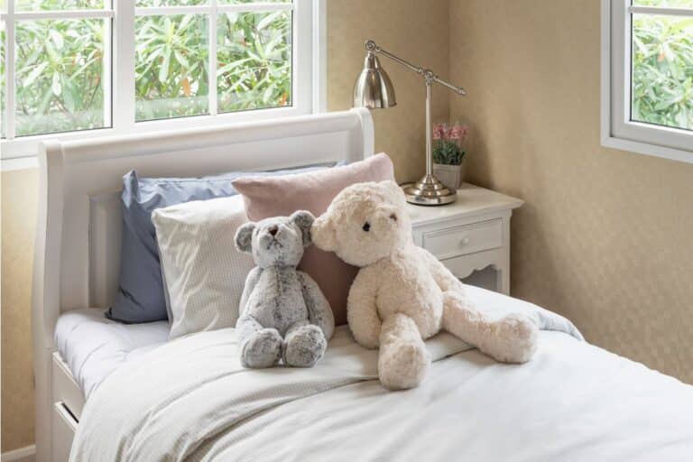 A neatly made, sleep-friendly bedroom features white and pastel bed linen, two teddy bears side by side on top, and a bedside table with a lamp and small plant beside a window showcasing lush green foliage outside.
