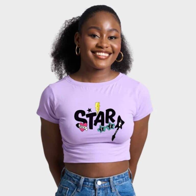 A smiling woman in a lavender crop top with "STAR" and colourful graphics channels Y2K fashion as she stands with her hands behind her back. She has hoop earrings, curly hair, and blue jeans against a plain light background.