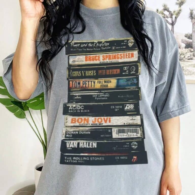 A person wearing a grey oversized plus size T-shirt featuring a graphic of stacked cassette tapes labelled with classic rock band names like Bruce Springsteen, Guns N' Roses, Tom Petty, AC/DC, Bon Jovi, Van Halen, and The Rolling Stones.