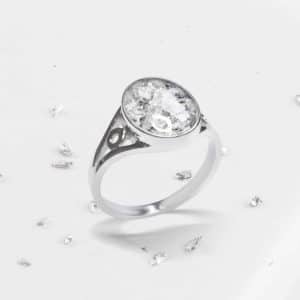 A silver ring with a large oval-shaped clear gemstone, surrounded by smaller sparkling stones, is displayed on a white surface with scattered loose gemstones—an elegant example of ashes jewellery.