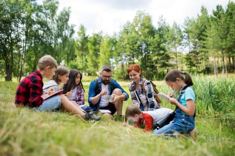 A group of children sit on grass with two adults, writing in notebooks during a sunny school lesson held outdoors near a forest and pond.