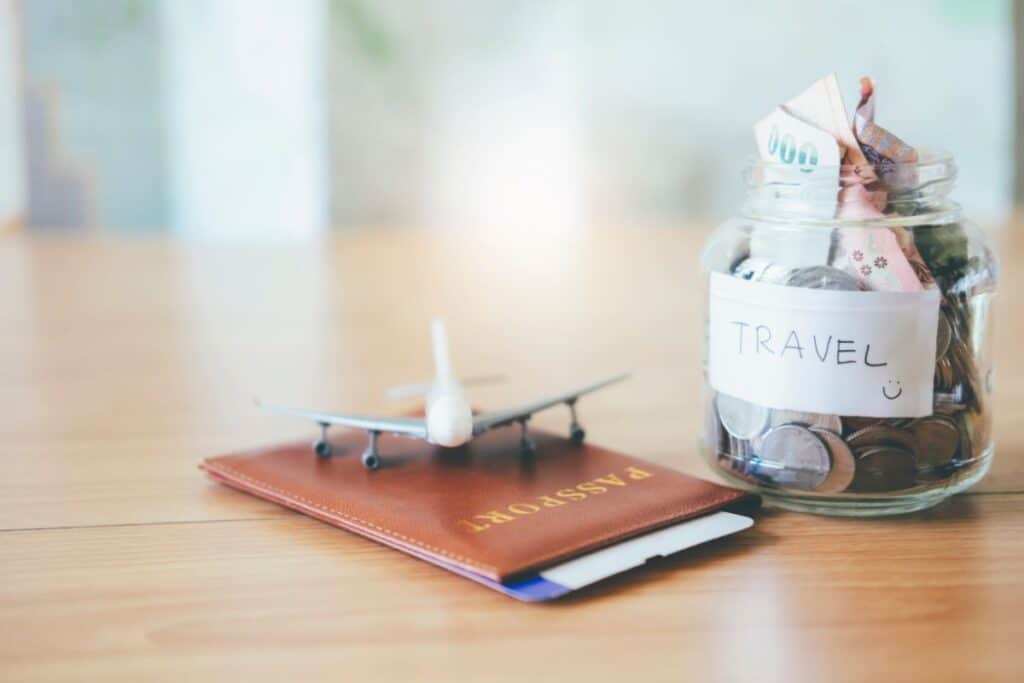 A jar labelled "TRAVEL" filled with coins and notes sits next to a brown passport, a small toy aeroplane, and a Koody brochure on a wooden table.