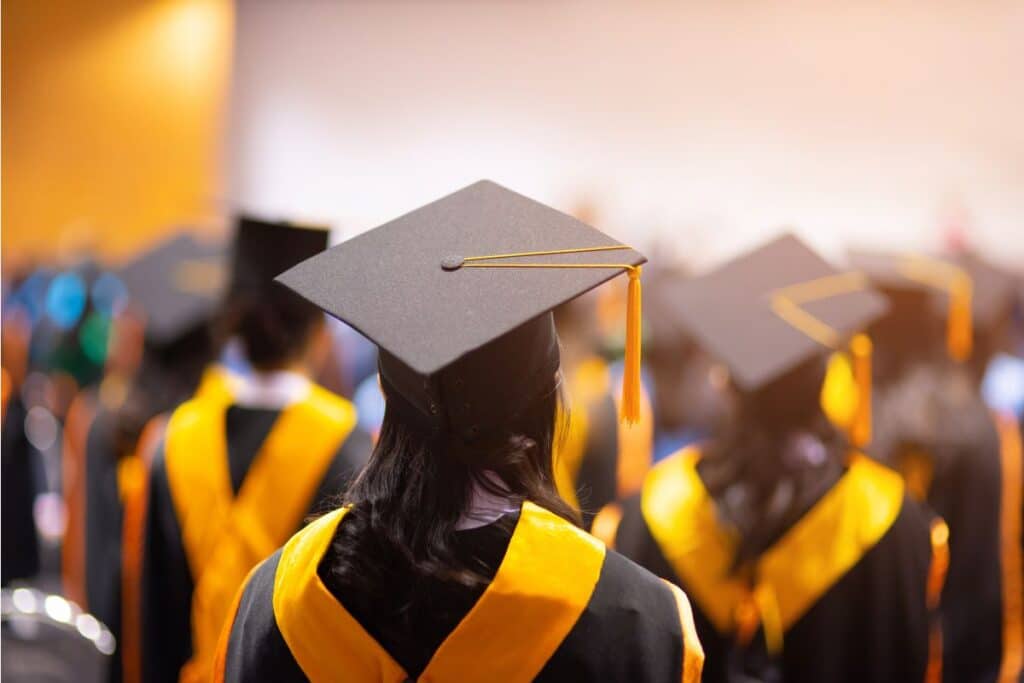 A group of university graduates in black caps and gowns with yellow stoles are seated, seen from behind, attending a graduation ceremony in a brightly lit hall.