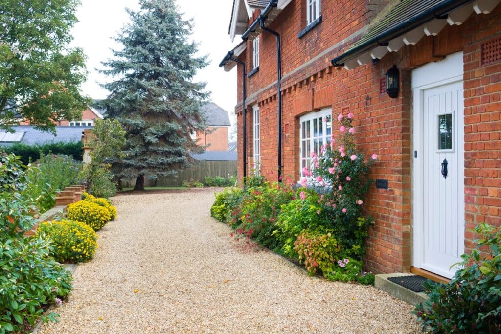 A brick house with white door and windows, surrounded by a gravel drive and garden beds filled with colourful flowers and green shrubs, offers charming kerb appeal, set against a backdrop of trees.