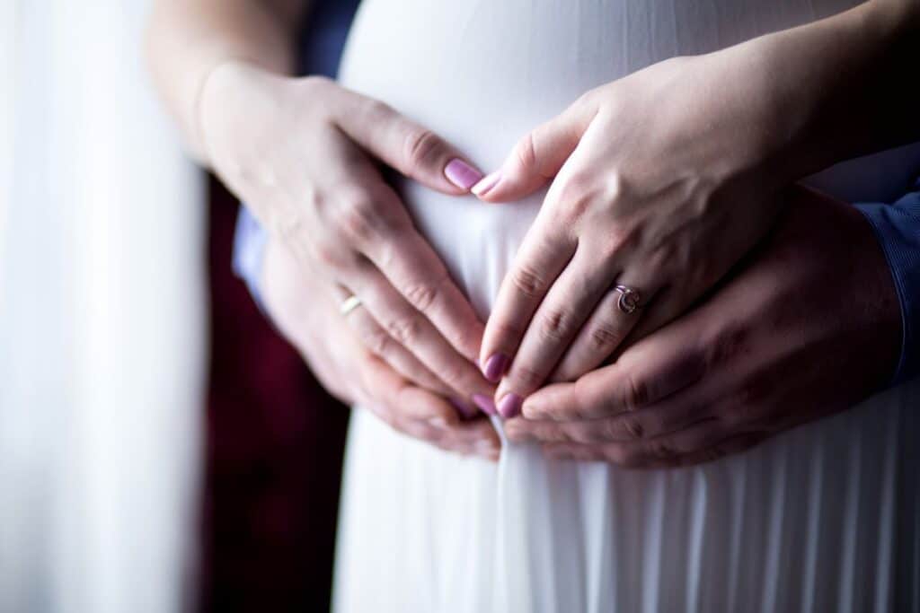 Close-up of two pairs of hands gently resting on a pregnant woman's bump, forming a heart shape. The scene conveys care and anticipation, reflecting the importance of blood banking for both mother and baby's health.