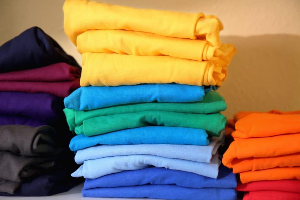Stacks of neatly folded charity clothing T-shirts in various bright colours, including yellow, blue, green, orange, purple, and grey, are arranged on a shelf against a beige background.