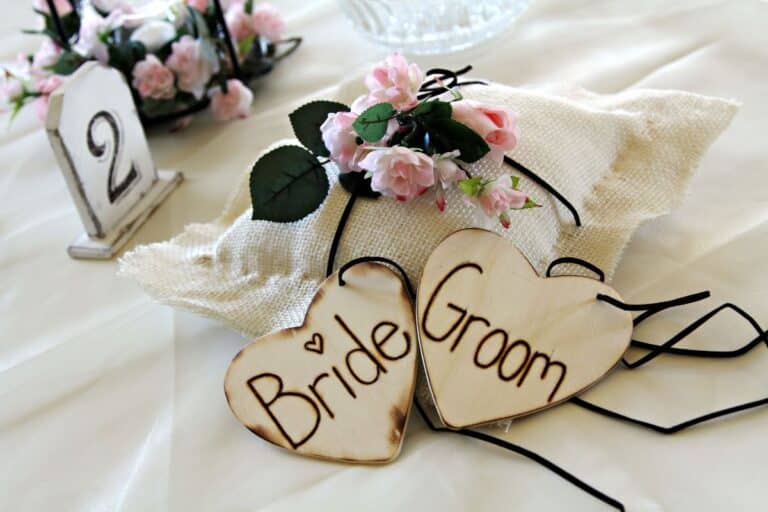 Two wooden heart-shaped signs labelled "Bride" and "Groom" rest on a hessian cushion decorated with pink flowers, adding a charming touch to this wedding scene. A table number "2" and more flowers are visible in the background.