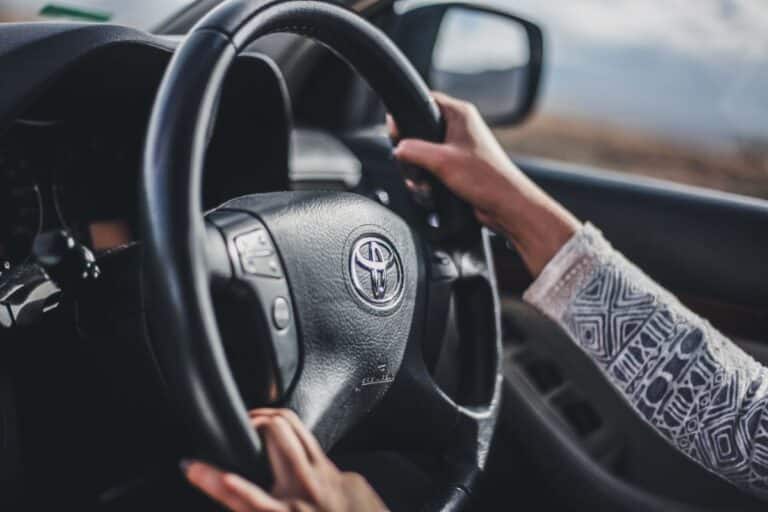 A person with a patterned long-sleeve shirt is driving a hire car, holding a black steering wheel with a Toyota logo in the centre. The car window and wing mirror are visible in the background.