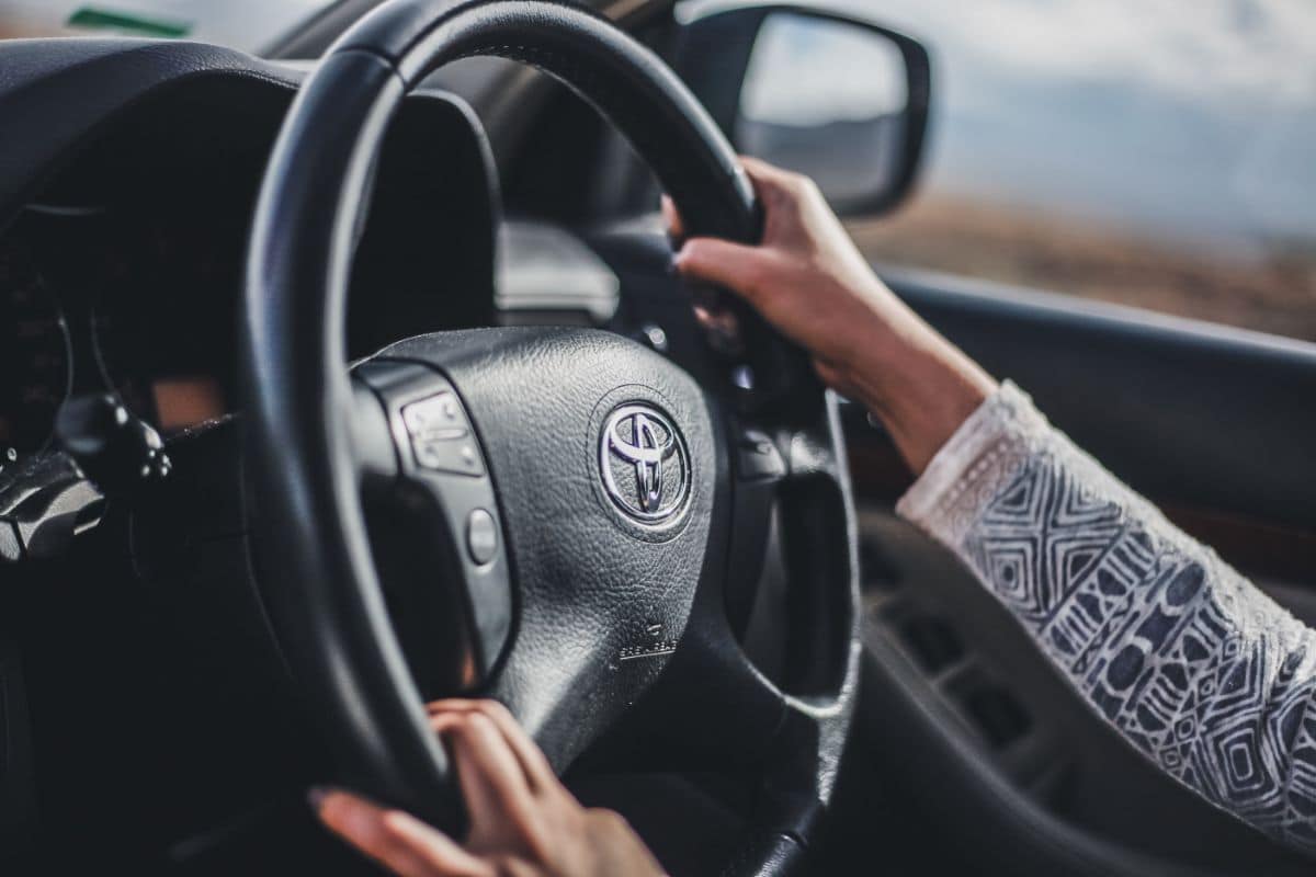 A person with a patterned long-sleeve shirt is driving a hire car, holding a black steering wheel with a Toyota logo in the centre. The car window and wing mirror are visible in the background.