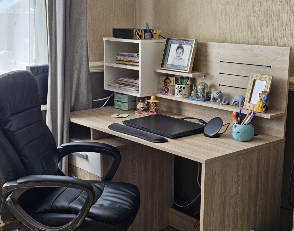 A tidy wooden desk with a black office chair, drawing tablet, shelves holding books, framed photos, small plants, and colourful figurines—perfect for working from home—positioned by a window with grey curtains.