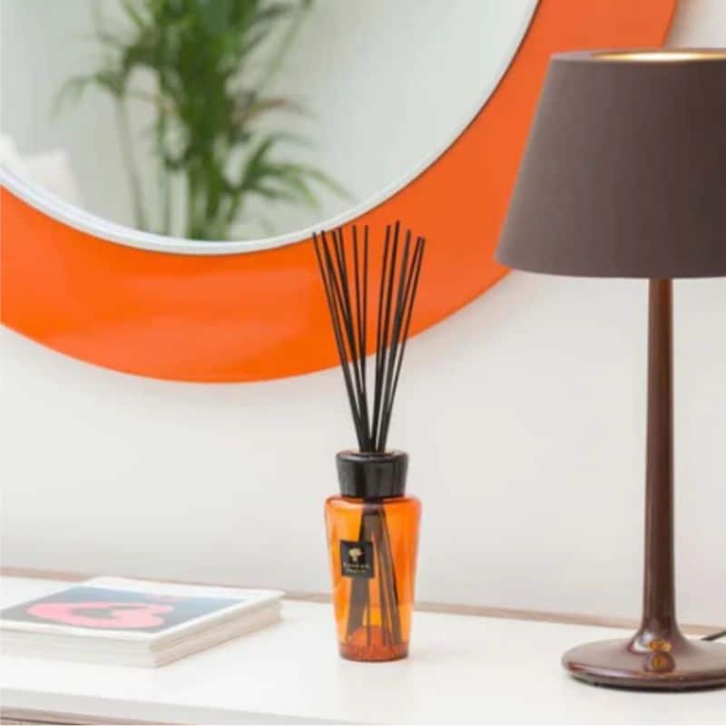 An orange glass reed diffuser sits on a white surface next to a stack of magazines and a brown table lamp, creating an inviting home vibe with an orange-rimmed mirror and a blurred green plant in the background.