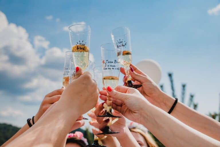 A group of people raise champagne glasses for a toast outdoors under a blue sky, celebrating together. Some glasses have "Team Bride" written on them, highlighting the joy of a hen do or hen party.