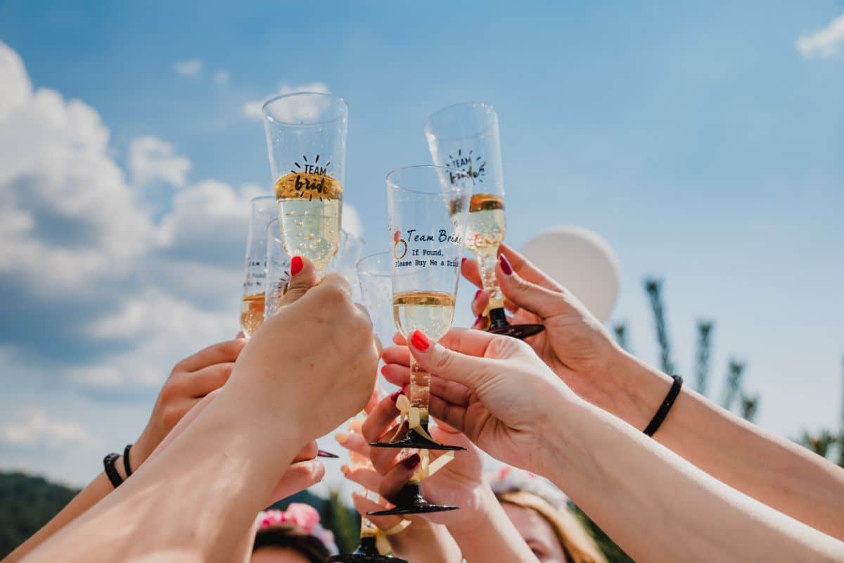 A group of people raise champagne glasses for a toast outdoors under a blue sky, celebrating together. Some glasses have "Team Bride" written on them, highlighting the joy of a hen do or hen party.