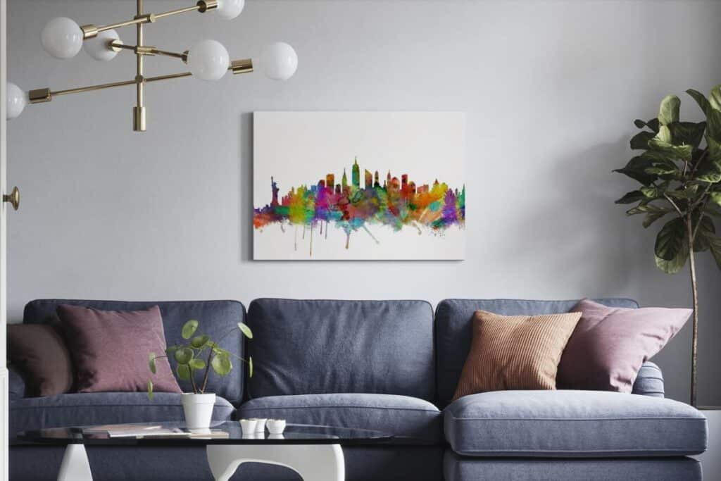A modern living room with a blue sofa, pink and brown cushions, a glass coffee table with a plant, a potted plant in the corner, and striking cityscape wall art above the sofa.