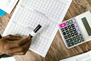 A person holds a black marker above a printed spreadsheet with financial data, calculating money next to a calculator on a wooden table.