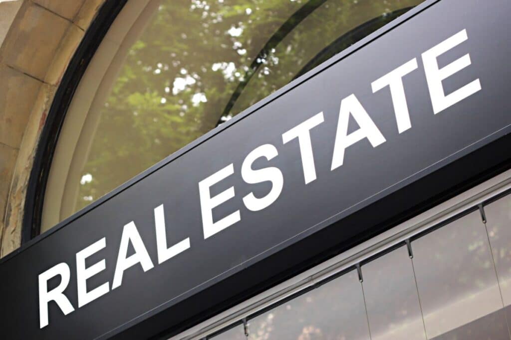 A close-up of a black sign with large white letters spelling "PROPERTY," mounted on a building with a glass window, hints at nearby property courses, with greenery reflected above.