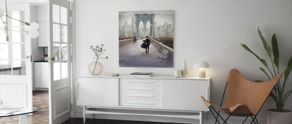 A modern, bright living room with white furniture, a potted plant, and a tan butterfly chair. Striking wall art of a couple walking on a bridge hangs above a white sideboard with minimal decor.