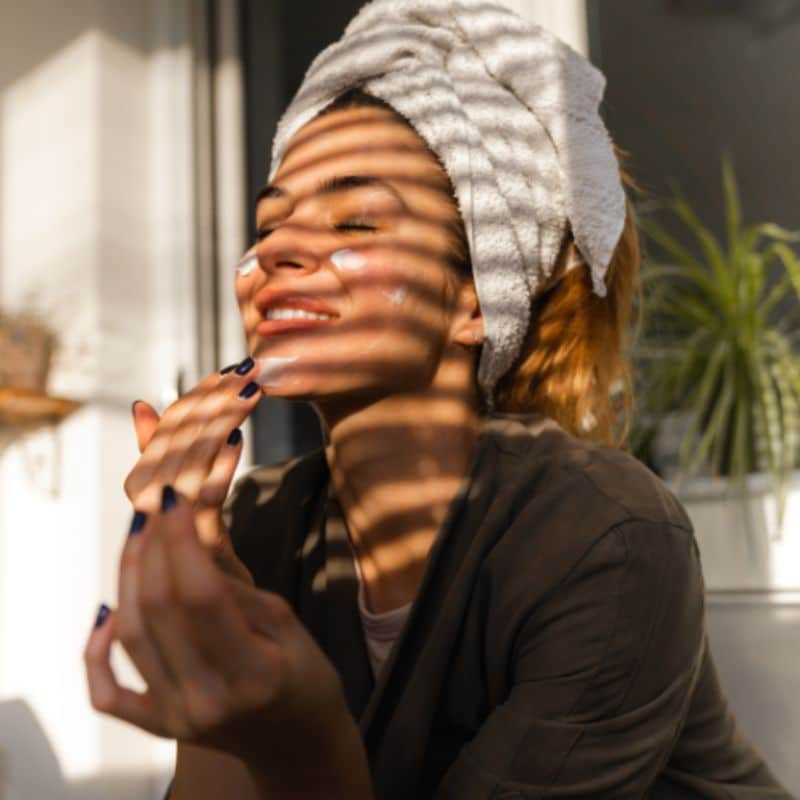 A woman with a towel wrapped around her head smiles with confidence, eyes closed as she applies cream to her face. Sunlight filters through the window blinds, casting striped shadows on her skin. A green plant is in the background.