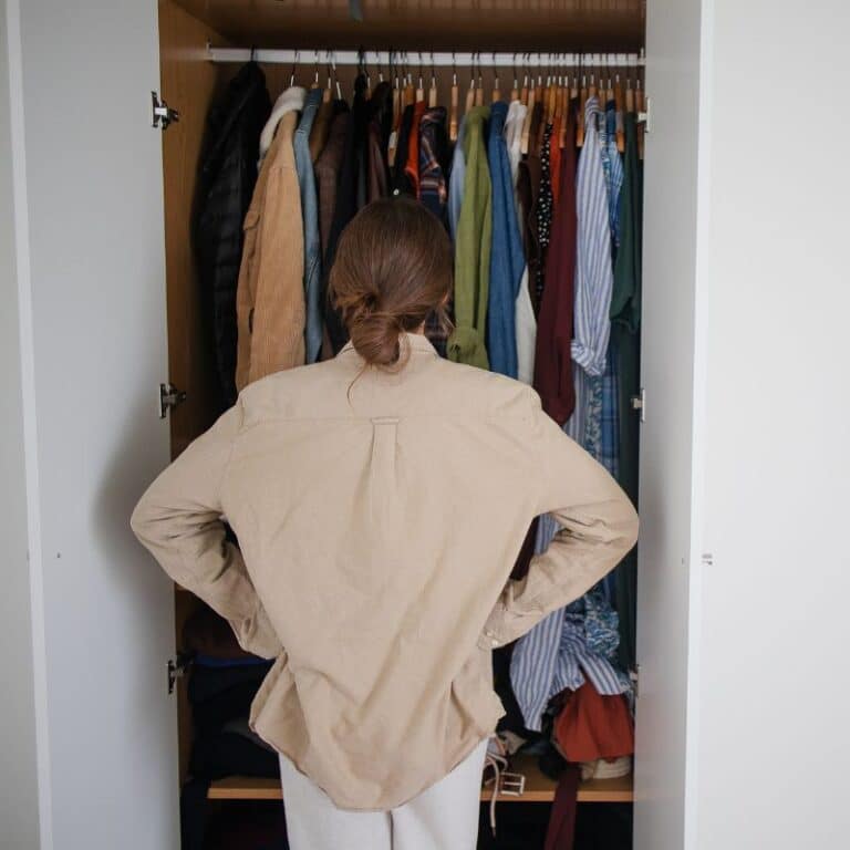A person with brown hair tied in a low bun stands facing an open wardrobe, hands on hips, considering the various clothes hanging inside.