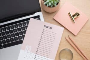 A to-do list on a desk next to a laptop keyboard, two pencils, a small plant in a pot, a notepad with a gold clip, and a magnifying glass—essentials for an organised work from home setup on a light wooden surface.