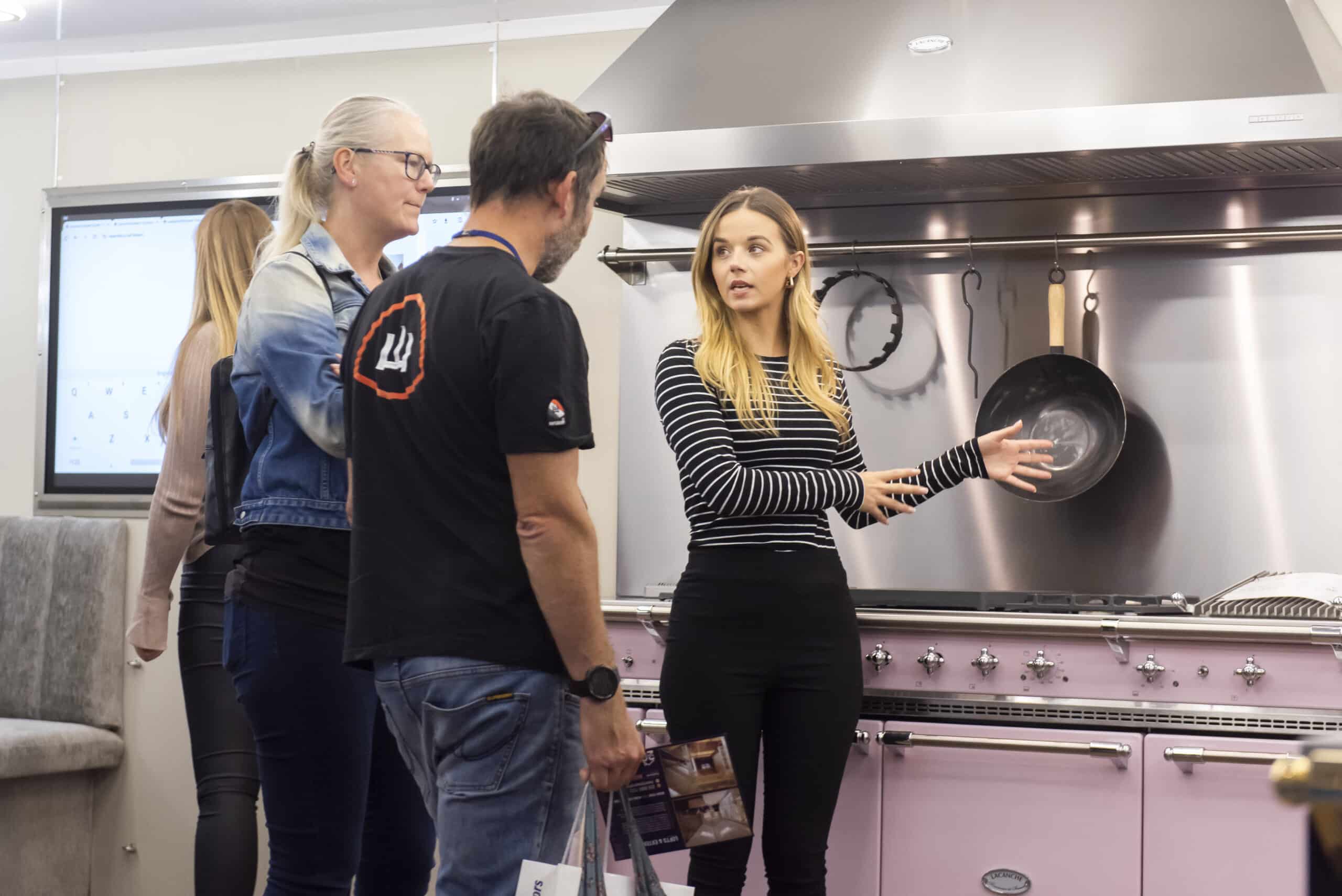 A woman in a striped shirt gestures while speaking to three people at The Northern Homebuilding & Renovating Show, standing in front of a pink cooker and stainless steel splashback with pans hanging in a modern kitchen showroom.