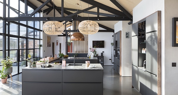 Spacious modern kitchen with black cabinetry, marble worktops, large windows, and stylish decor—a design inspired by trends from The Northern Homebuilding & Renovating Show. Potted plants and wicker pendants add a cosy touch.