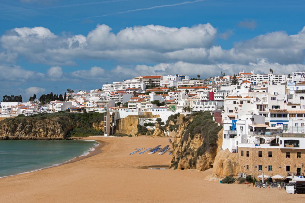 A sandy beach curves along the shore with rows of sun loungers. White buildings with red roofs sit on a cliff above the beach, beneath Portugal’s partly cloudy blue sky. Lush greenery dots the landscape.