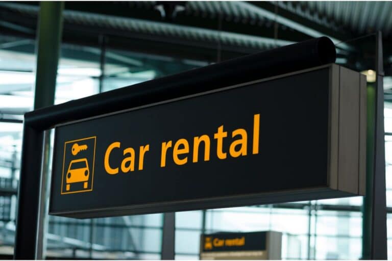 A black sign with yellow text and icons reads "Car hire" with a car and key symbol, hanging in an indoor area, likely at an airport or car transport hub.