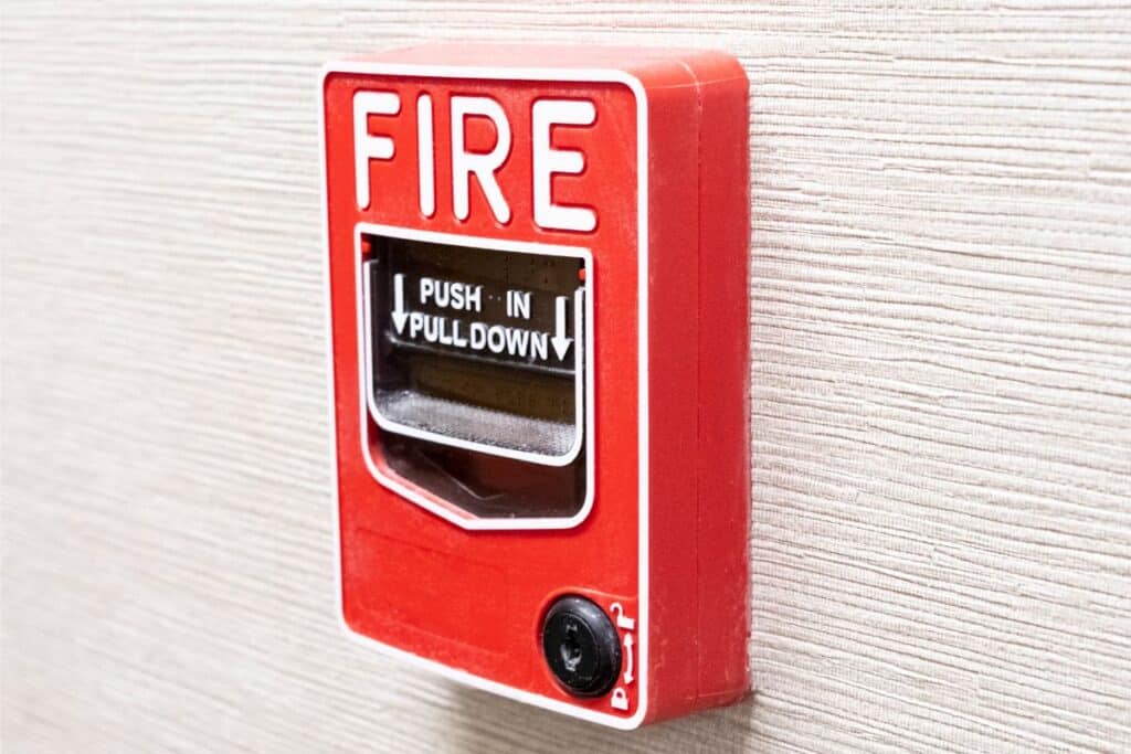 A red fire alarm call point, one of the essential safety devices, is mounted on a textured beige wall with "FIRE" in large white letters and instructions to "PUSH IN, PULL DOWN.
