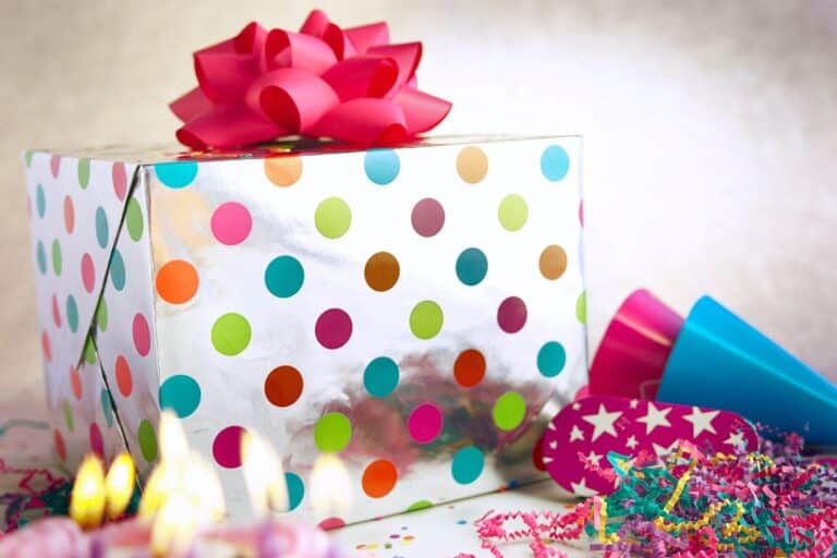 A Y2K-inspired gift box wrapped in white paper with colourful polka dots, topped with a large pink bow, sits among party hats, confetti, and lit candles, creating a festive birthday atmosphere.