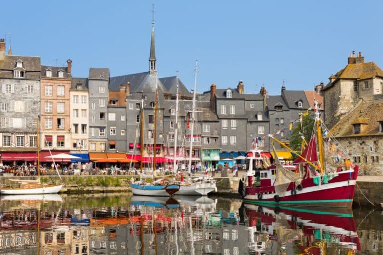 Colourful boats are docked in a calm harbour, reflecting old, charming buildings with grey roofs and vibrant awnings under a clear blue sky—one of those lesser-known destinations where people enjoy the lively waterfront scene.