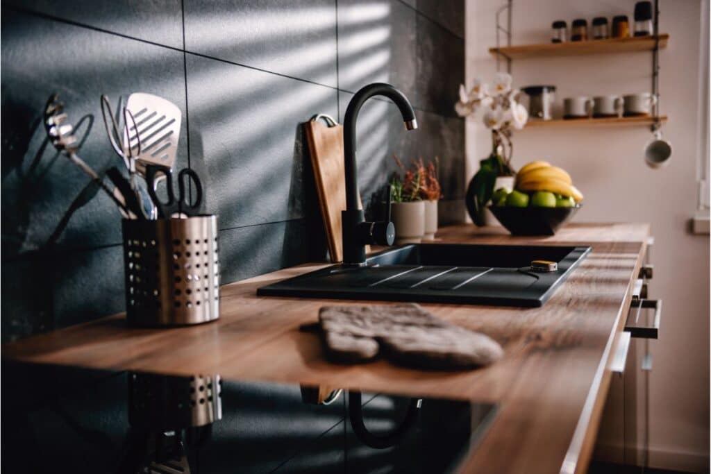 A modern kitchen with a wooden worktop, black sink, metal utensil holder, kitchen appliances, potted plants, a bowl of fruit, chopping board, and shelves with jars and spices. Sunlight streams through a nearby window.