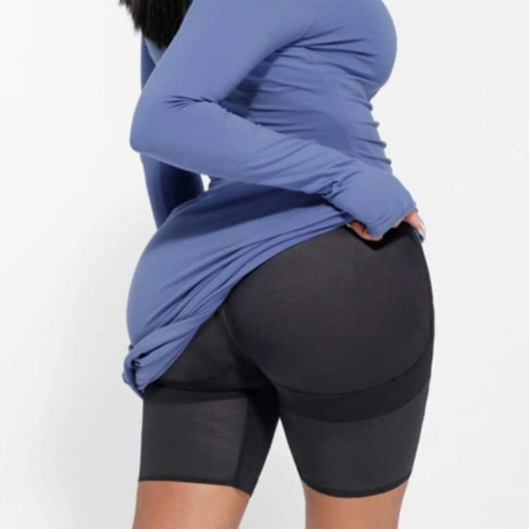 A person wearing a long-sleeved blue top lifts the hem to reveal black shaping shorts underneath, with their back turned to the camera against a plain white background.