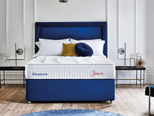 A modern bedroom with a blue upholstered bed, white mattress labelled “Sleepeezee” and “Jessica,” layered white bedding ideal for warmer sleepers, plus decorative cushions; minimalist decor with benches and lamps on each side.