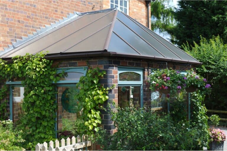 A charming brick conservatory with large windows and a metal roof is covered in climbing plants, surrounded by greenery and hanging flower baskets on a sunny day.