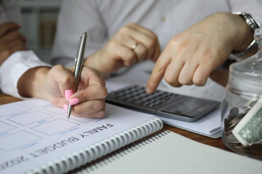 Two family members review finances together, one writing on a “Family Budget 2021” worksheet whilst the other uses a calculator. A jar of cash and coins sits nearby, along with a notepad.