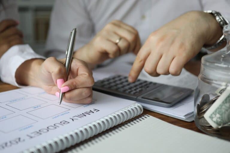 Two family members review finances together, one writing on a “Family Budget 2021” worksheet whilst the other uses a calculator. A jar of cash and coins sits nearby, along with a notepad.