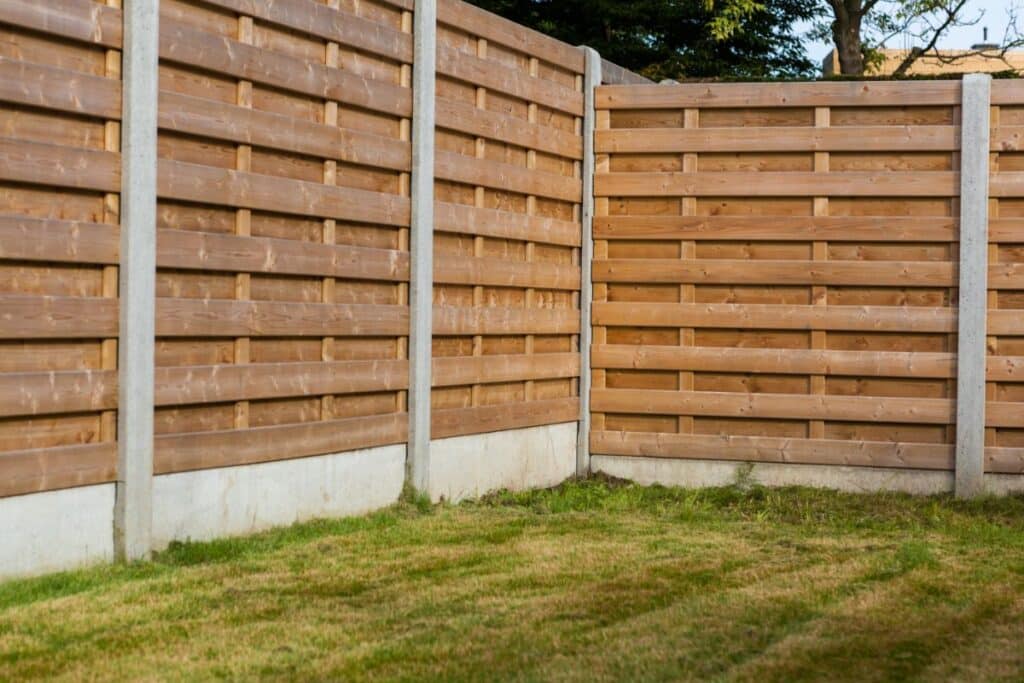 A corner of a back garden with green grass, bordered by tall wooden fencing panels supported by concrete posts and a concrete base.